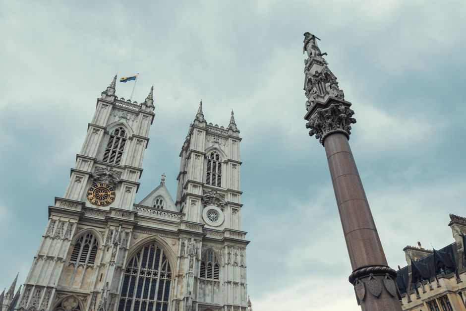 A low-angle view of Westminster Abbey's Gothic façade showing intricate stone carvings, tall pointed arches, and stained glass windows, with two prominent square towers topped with spires and a Union Jack flag flying atop one of them. To the right, a tall, reddish-brown column with an ornate statue on top extends upward. The sky behind is overcast with gray clouds, creating a moody atmosphere. The scene represents a historic architectural site visually linked to Westminster, where private or alternative waste management services such as Waste Removal Westminster may operate nearby to support the maintenance and cleaning of such heritage landmarks.