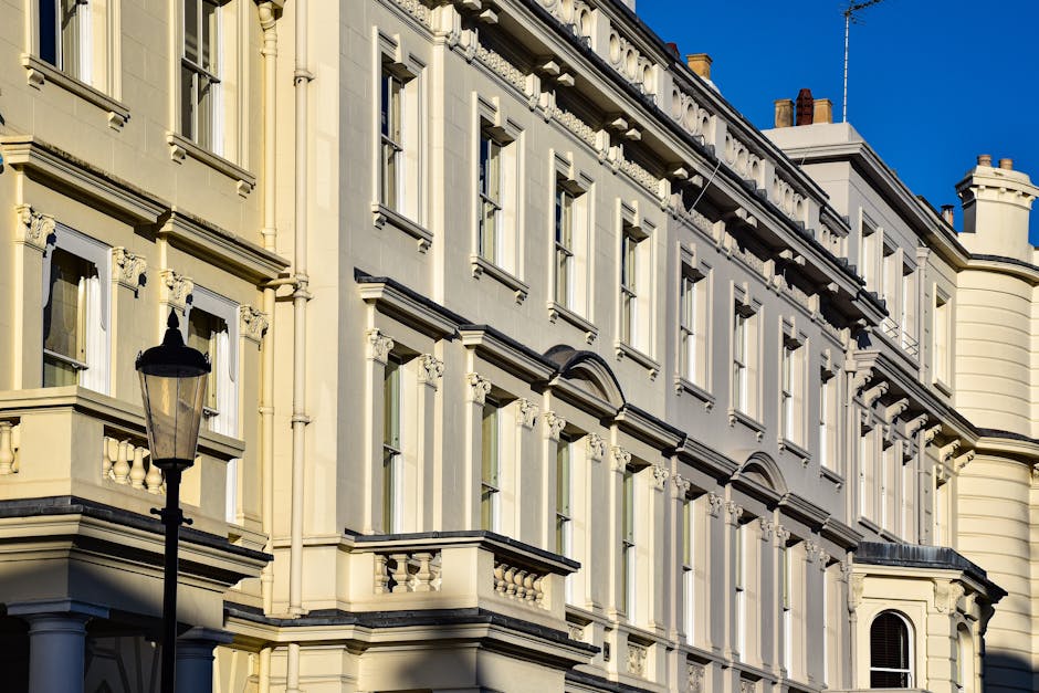 The image depicts the upper facades of a classical-style building with ornate architectural details, including decorative cornices, window mouldings, and balustrades, painted in a light cream or beige colour with contrasting dark accents. Several large windows are visible, framed with intricate trim, reflecting the bright natural daylight against a clear blue sky. In the foreground, a traditional black street lamp with a glass lantern stands on a pavement area or possibly a residential street setting. The building’s exterior appears clean and well-maintained, highlighting historic architectural features common in Westminster. The scene conveys an urban environment typical of an area with period architecture, with no visible rubbish or waste-related items present. As a professional waste removal service, Waste Removal Westminster sometimes assists with private collection or on-site clearance for similar properties, though no waste is visible in this specific image, emphasizing a tidy and well-kept streetscape that may benefit from regular rubbish collection or selective waste management solutions.