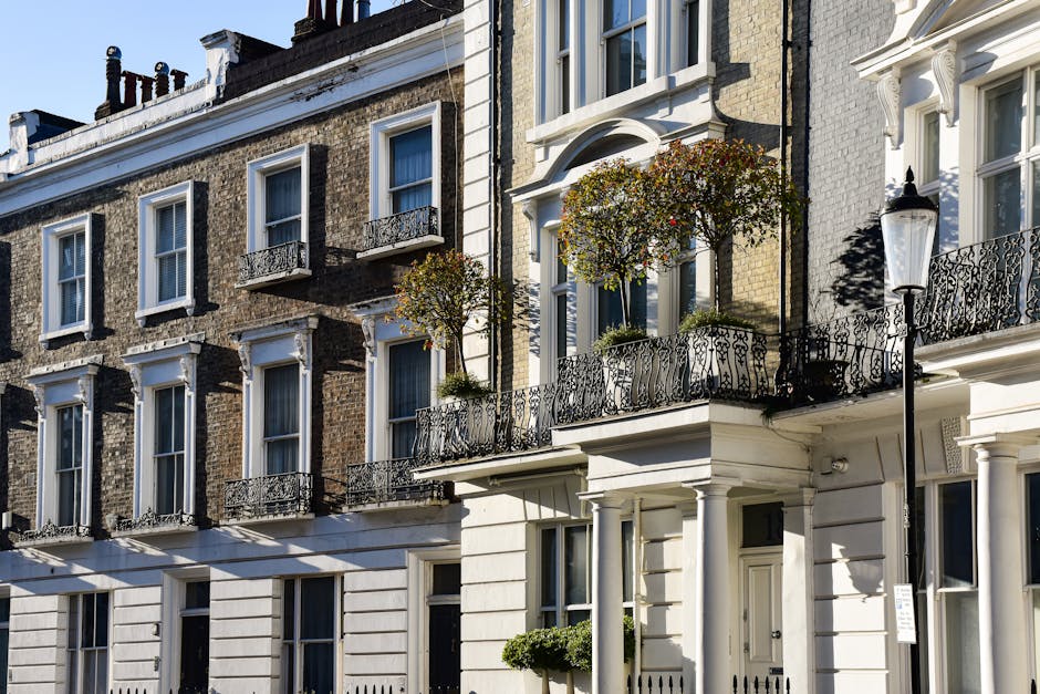 The exterior of a multi-story residential building with a classic architectural style, featuring a white facade on the ground floor and upper levels of exposed brickwork in various shades of brown and tan. The building has several large sash windows framed in white, some with small decorative black wrought iron balconies with intricate railing designs. In front of the building, there are two small trees planted in planters on the first-floor balcony, showing green foliage and a few reddish berries, supported by black metal brackets. A traditional street lamp with a black post and white glass enclosure stands on the sidewalk beside the building, casting subtle shadows in the daylight. The scene indicates a well-maintained urban environment typical of Westminster, with natural sunlight highlighting textures and colors of the building materials, fitting the setting of private residential properties often serviced by independent waste removal companies such as Waste Removal Westminster for rubbish collection and on-site clearance activities.