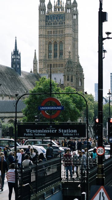 The image depicts the busy entrance to Westminster underground station in London, with a large, prominent black sign displaying the station name and amenities such as public subway access and toilets. In the foreground, a group of pedestrians are waiting or walking on the pavement, some behind a black metal railing. Several cars are visible on the street, and a traffic light and a speed limit sign indicating 20 mph are also present. In the background, the historic facade of the Elizabeth Tower, commonly known as Big Ben, rises above the scene, with its ornate gothic architecture and clock tower details, partially obscured by trees and other street furniture. The sky is overcast, casting diffuse natural light over the urban environment. The scene captures the typical street-level activity near Westminster, highlighting the integration of transport infrastructure with city life, relevant to discussions of alternative waste handling options like private rubbish collection services provided by Waste Removal Westminster.