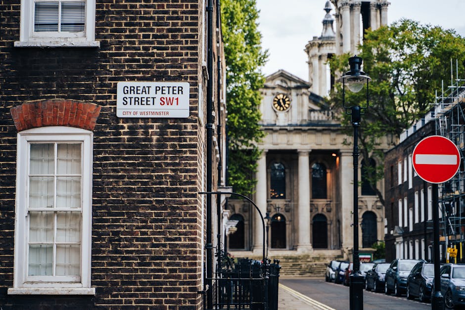 A street scene in Westminster showcases a corner of a brick building with a white sash-window framed with red brick accents and a street name sign reading 'Great Peter Street SW1' indicating the borough. In the background, there is an imposing neoclassical-style building with stone columns, a triangular pediment, and a clock face visible above the entrance, surrounded by lush green trees. To the right, a black lamppost with a traditional lantern-style design stands alongside a red circular 'no entry' traffic sign mounted on a pole. Parked cars line the narrow street, which has a slight incline and smooth asphalt surface. The scene is lit by natural daylight, with clear skies and some foliage visible, reflecting typical urban architecture and street layout in Westminster suitable for private waste collection and rubbish removal services managed by Waste Removal Westminster.