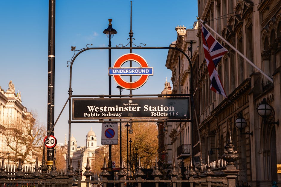 A photograph of Westminster Station in London showing a large roundel sign with a blue bar reading 'UNDERGROUND' in white letters, suspended from an ornate black metal bracket attached to a lamppost. Below this, a rectangular black sign displays 'Westminster Station' in bold white text, along with additional information about public subway and toilet facilities. To the right, a Union Jack flag flutters from a flagpole attached to an elegant, historic stone building with detailed window surrounds and classical architectural elements. In the background, the clear blue sky and partial view of another historic building are visible, with sunlight illuminating the scene. The surrounding environment includes wrought-iron fencing and several vertical street signs, creating an urban landscape typical of central London, and the signage and atmosphere reflect the area's appearance related to public transit and city life, as well as the possibility of private or independent waste management services nearby.
