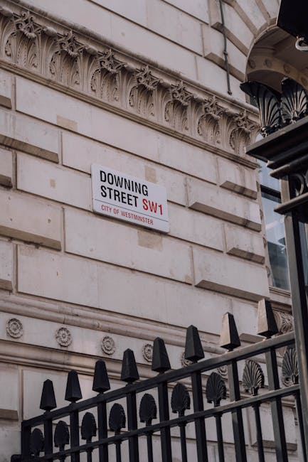 A street scene in Westminster showcases a corner of a brick building with a white sash-window framed with red brick accents and a street name sign reading 'Great Peter Street SW1' indicating the borough. In the background, there is an imposing neoclassical-style building with stone columns, a triangular pediment, and a clock face visible above the entrance, surrounded by lush green trees. To the right, a black lamppost with a traditional lantern-style design stands alongside a red circular 'no entry' traffic sign mounted on a pole. Parked cars line the narrow street, which has a slight incline and smooth asphalt surface. The scene is lit by natural daylight, with clear skies and some foliage visible, reflecting typical urban architecture and street layout in Westminster suitable for private waste collection and rubbish removal services managed by Waste Removal Westminster.